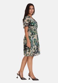 Robe courte à manches courtes à motifs floraux dans des teintes de vert et de beige, avec une taille élastique. Associée à des mules noires à talons avec un bout pointu.