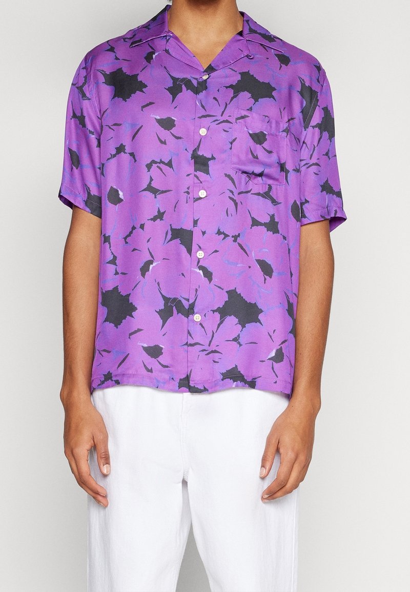 Homme portant une chemise violette à manches courtes avec un motif floral noir et un pantalon blanc, debout devant un fond uni.