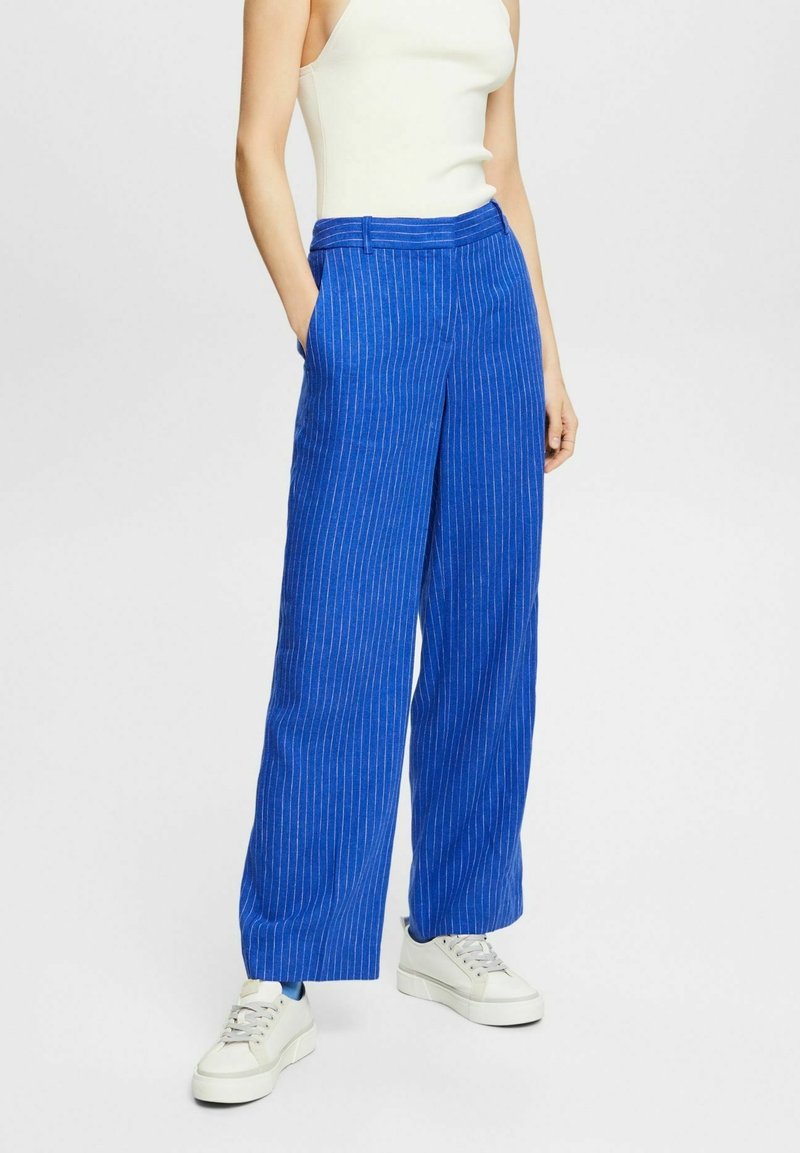 Femme portant un pantalon large à rayures bleues, un haut blanc sans manches et des baskets blanches, debout avec une main dans la poche.