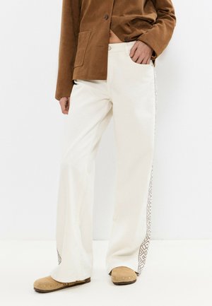 Personne portant un pantalon blanc à jambes larges avec des bandes latérales à motifs, des sabots en daim bronzé cloutés, et une veste marron avec une poche avant.