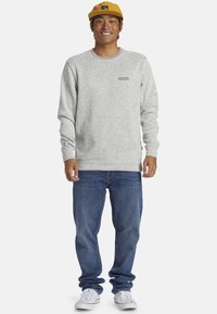 Grauer Fleece-Sweatshirt mit gerippten Bündchen und Saum, kombiniert mit blauen Jeans und weißen Sneakers. Gelbe Kappe mit Logo-Akzent.