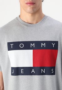 Hallhalli puuvillane t-särk, millel on graafiline print tekstiga "TOMMY" "JEANS" peale valgete ja tumesiniste tähtedega punase ja valge ploki taustal.
