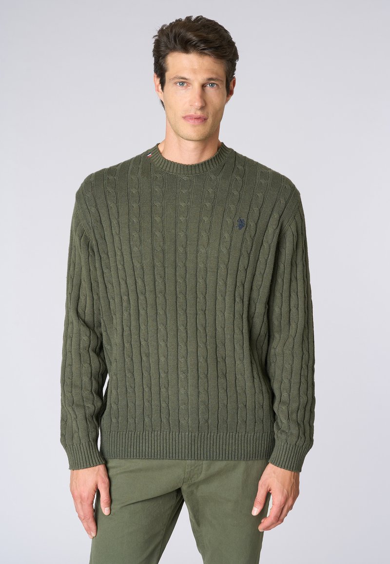 Maglione a girocollo in maglia cable-knit verde scuro, con polsini e orlo a costine. Presenta un piccolo logo sul petto. Indossato con pantaloni oliva coordinati.