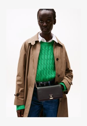 Donkerharige vrouw die een beige trenchcoat draagt over een groene kabelgebreide trui, witte blouse, blauwe spijkerbroek en een zwarte crossbodytas.