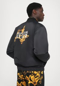 Man som bär en svart bomberjacka med guld barockdesign och texten "Versace Jeans Couture", i kombination med matchande mönstrade byxor.