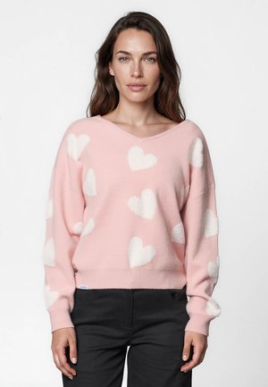 Femme aux longs cheveux bruns portant un pull rose à motifs de cœurs blancs et un pantalon noir sur un fond blanc uni.