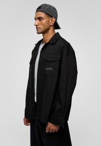 Zwarte button-up jas met twee borstzakken en tekstdetails. Gedragen over een grijs shirt. Model draagt een grijze pet. Van katoen, met een ontspannen pasvorm.