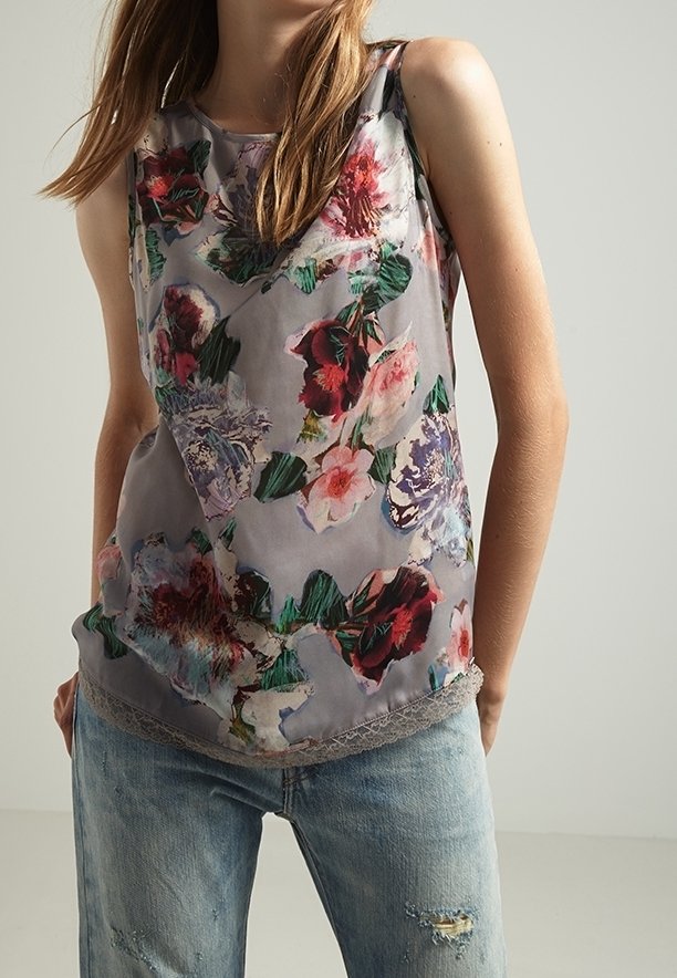 Haut sans manches à imprimé floral avec des fleurs roses, rouges et vertes sur fond gris, porté avec un jean bleu clair délavé.