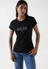 Schwarzes Baumwoll-T-Shirt mit einem mit Strass verzierten "SALSA"-Logo und "JEANS" in Blau, kombiniert mit blauen Jeans. Kurze Ärmel.