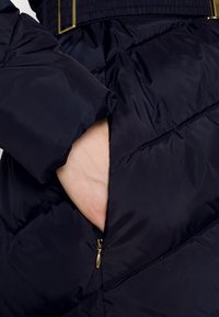 Tmavě modrá prošívaná bunda s diamantovou texturou prošívání, s rukou v boční kapse na zip a detaily zlatých zipů.