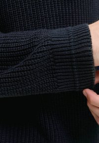 Sweter dzianinowy w ciemnym granacie, z wyraźnym ribbrowym wzorem. Zbliżenie na mankiet, podkreślające grubość materiału i szczegóły ściegów.