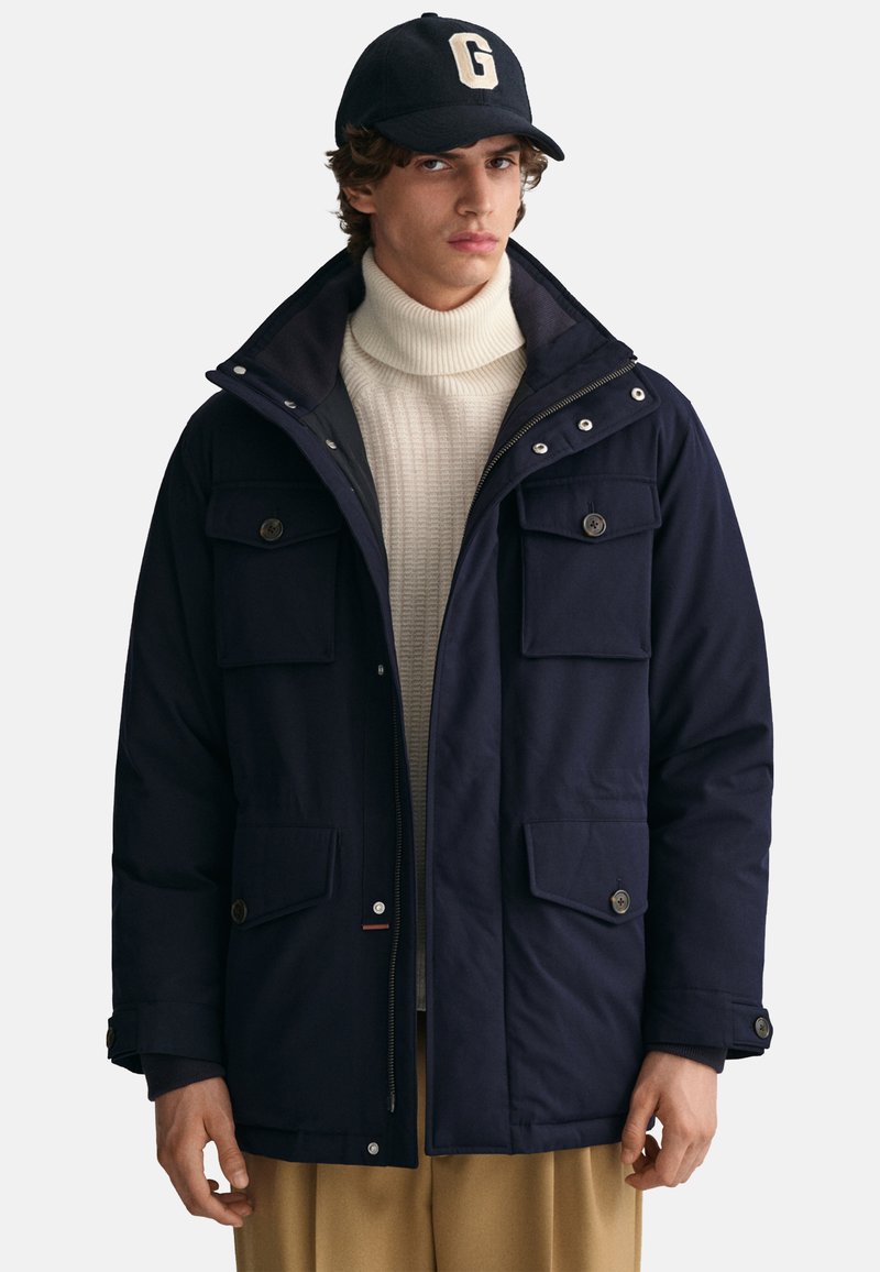 Giovane uomo che indossa una giacca blu navy, un maglione colletto alto crema, pantaloni color cammello e un cappellino nero con una grande "G" bianca sulla parte frontale.