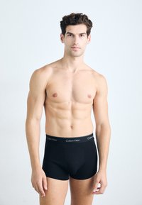 Boxeri scurți din bumbac negru, cu un tiv elastic branduit. Design ajustat afișat pe un model cu o construcție musculară.