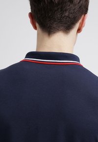 Granatowa koszulka polo z teksturowanym wykończeniem, z kontrastowymi czerwonymi i białymi detalami na kołnierzu. Z tyłu widać czysty, prosty design.