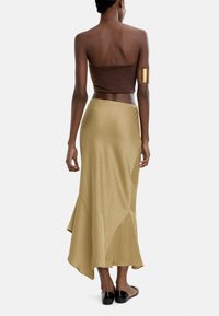 Brązowy top bez ramiączek w połączeniu z zwiewną złotą spódnicą o asymetrycznym hemie, gładkiej teksturze i delikatnym połysku. Czarne płaskie buty.