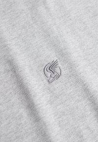 Graues Stoff-T-Shirt mit weicher Textur und subtiler Melierung. Verfügt über ein in dunklerem Grau eingesticktes Logo.