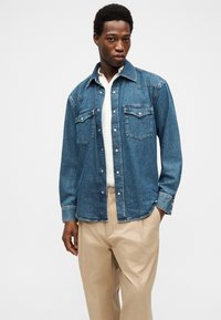 Άνδρας με μπλε πουκάμισο τζιν κουμπωτό πάνω από λευκό τοπ και μπεζ παντελόνι, στέκεται με ένα χέρι στην τσέπη του μπροστά σε λευκό φόντο.