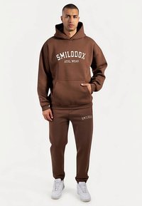 Ensemble sweat à capuche marron et pantalon de survêtement avec texte blanc "SMILODOX ATHL WEAR". Tissu épais, poche kangourou et poignets élastiques. Baskets blanches.