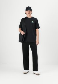 T-shirt en coton noir avec logo vert, pantalon noir à coupe droite, casquette noire et sac fourre-tout noir mat. Baskets blanches avec des accents beiges.