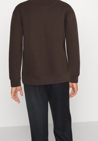 Sudadera marrón de cuello redondo con mangas largas, puños y dobladillo acanalados. Textura suave, costuras mínimamente visibles, combinada con pantalones oscuros.