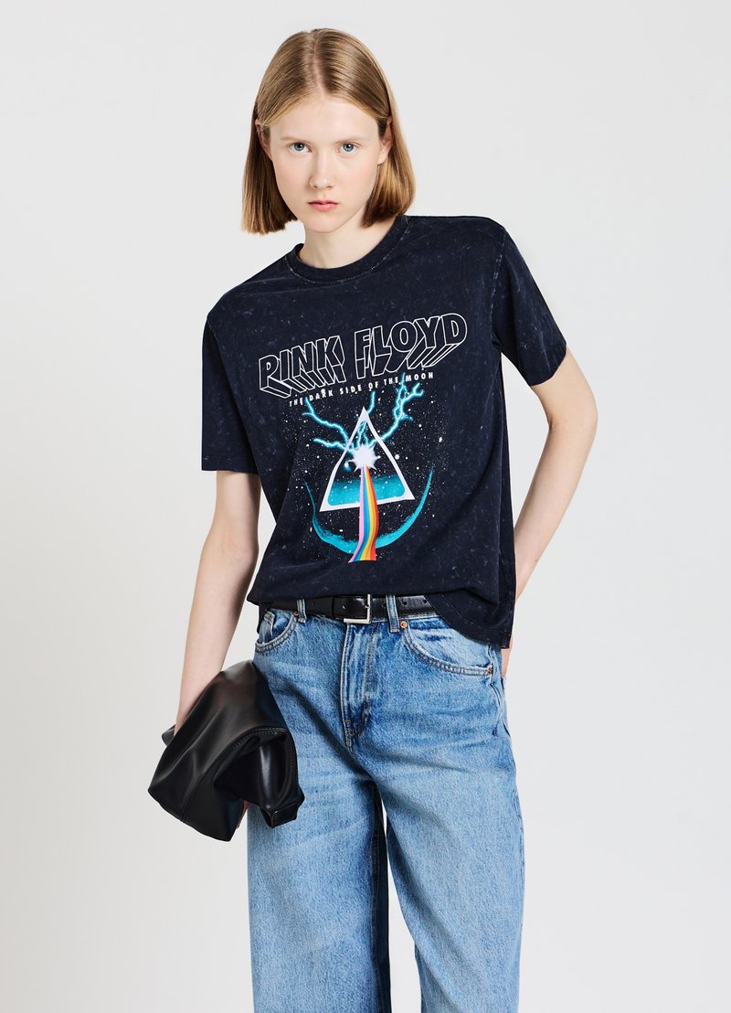 T-shirt nera in cotone con grafica "Pink Floyd", design di prisma e arcoiris, maniche corte, vestibilità rilassata, indossata con jeans azzurri chiari.