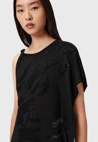 Nuori nainen, jolla on suorat tummat hiukset, on pukeutunut mustaan epäsymmetriseen yläosaan, jossa on kiiltäviä mustia helmia koristeluina ja solmittu yksityiskohta olkapäässä.