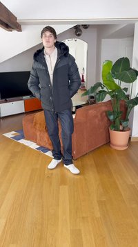 Svart pufferjacka med pälsfodrad huva, grå skjorta, mörka jeans och vita sneakers. Står i ett rum med trägolv och växter.