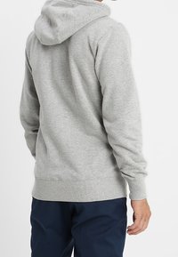 Sudadera gris con capucha y mangas largas, tejido texturizado, puños y dobladillo acanalados, y un bolsillo tipo canguro. Vista trasera, combinada con pantalones oscuros.