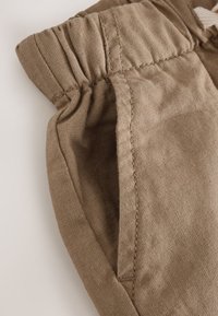 Detailní záběr kapsy béžových kalhot s našitým elastickým pasem a viditelnými detaily stehů.