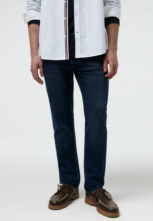 Mężczyzna ubrany w ciemnoniebieskie dżinsy, brązowe skórzane buty sznurowane oraz białą koszulę zapinaną na guziki z podwiniętymi rękawami na jednolitym tle.