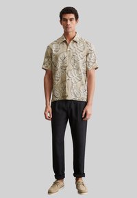 Kurzarmhemd aus beigem Stoff mit schwarzen Blattmustern; kombiniert mit schwarzen Hosen und beigen Schuhen. Einfaches Button-Down-Design.
