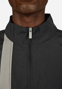 Μαύρο μπουφάν με φερμουάρ και ψηλό γιακά, που διαθέτει μια μπεζ ρίγα στο μπροστινό μέρος και ασημένιο φερμουάρ με λεπτομέρειες της μάρκας.