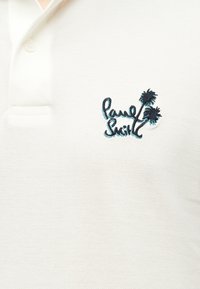 Camisa tipo polo blanca con un cuello abotonado y un logo oscuro bordado con dos palmeras y texto cursivo en el pecho.