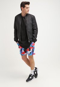 Chaqueta bomber negra, camiseta negra, bañador multicolor con patrón geométrico, zapatillas negras con acentos blancos, calcetines blancos.