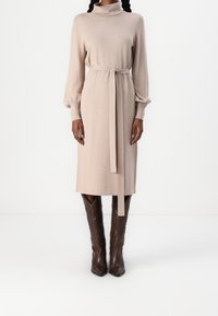 Vestito beige lavorato a maglia con maniche lunghe e gonfie e cintura in vita. Abbinato a stivali cowboy in pelle marrone con dettagli intricati.