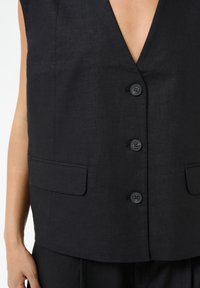 Črno brezrokavno telovnik z globokim V-izrezom, ki ima štiri sprednje gumbe in dva stranska žepa, narejen iz teksturirane tkanine.