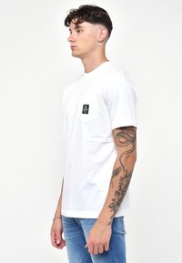 T-shirt bianco in cotone con scollatura rotonda e taschino a sinistra con un dettaglio grafico nero. Modello mostrato che indossa jeans blu.