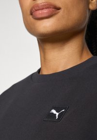 Camiseta de algodón negra con un parche cuadrado negro que presenta un logo blanco de Puma, cuello redondo y mangas cortas. Textura suave.