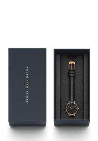 Zwart leren horlogeband met roségouden accenten in een marineblauwe doos. Rond horlogekastje met minimalistisch ontwerp en gouden markers, voorzien van een gleufgesp.