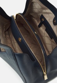 Marinblå handväska i nubuckläder med öppen topp, guldzipperficka och beige tygfoder tryckt med kedjor och varumärkeslogotyper.