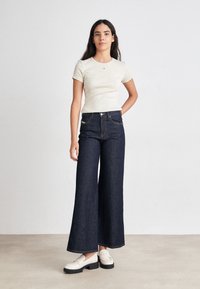 Lys beige tettskjorte med korte ermer, kombinert med vide mørk denim jeans. Modellen har på seg hvite loafers med en kraftig svart såle.