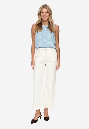 Femeie blondă purtând un top fără mâneci brodat, albastru deschis, pantaloni albi largi, cu talie înaltă, și pantofi negri cu toc ascuțit, stând în picioare și zâmbind.