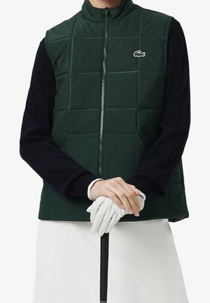 Person trägt eine dunkelgrüne gesteppte Weste über einem schwarzen Langarmshirt und hält einen weißen Golfhandschuh am Griff eines Golfschlägers.