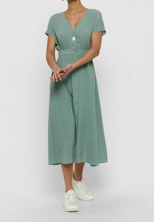 Femme portant une robe midi boutonnée vert sauge à manches courtes et des baskets blanches, se tenant les mains croisées devant.