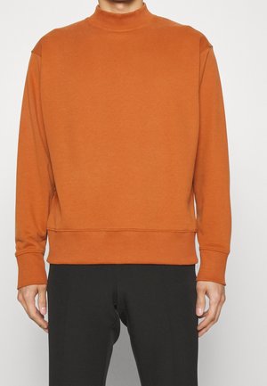 Homme portant un sweat-shirt uni orange brûlé à manches longues, associé à un pantalon noir, debout devant un fond blanc.