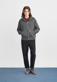 Sudadera gris con cremallera y bolsillo frontal, combinada con pantalones negros y zapatillas de senderismo grises. Tejido suave con cordones y detalle de logo.