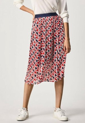 Femme portant une jupe midi plissée aux motifs abstraits rouges, blancs et bleu marine, associée à des baskets blanches et un pull blanc texturé.
