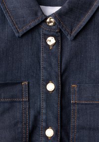 Chemise en denim bleu foncé avec surpiqûres orange, col pointu, deux poches poitrine et quatre boutons-pression métalliques au centre.