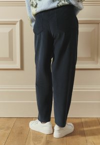 Tmavomodré manšestrové nohavice s voľným strihom a dvoma zadnými vreckami, kombinované s bielymi teniskami na drevenej podlahe.