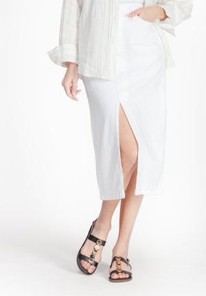 White midi skirt with a high slit, made of lightweight fabric. Paired with black sandals featuring circular gold hardware accents.